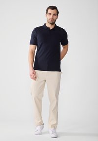 Polo din bumbac cu dungi navy, cu mâneci scurte, guler cu două butoane, asortat cu pantaloni bej și adidași albi. Textură netedă, design casual.