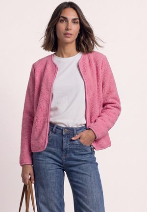 Femme portant une veste polaire rose sur un t-shirt blanc et un jean bleu, tenant un sac à main marron avec une main dans la poche.