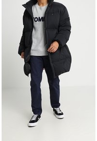 Persona che indossa un lungo cappotto invernale nero, un maglione grigio con testo blu navy, pantaloni blu navy e scarpe da ginnastica bianco e nero, in piedi su uno sfondo chiaro.