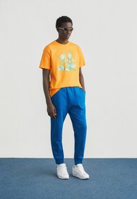 Πορτοκαλί βαμβακερό t-shirt με πράσινο γραφικό σχέδιο σε συνδυασμό με μπλε παντελόνια. Λευκά αθλητικά παπούτσια. Απλά γυαλιά ηλίου. Απλό φόντο.