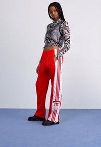 As calças de treino vermelhas apresentam riscas brancas laterais com botões de pressão e um logotipo da Adidas. A blusa preta de mangas longas tem um padrão de zebra e lantejoulas.
