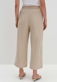 Beige-weite Hosen mit einem gerafften elastischen Bund, ausgestattet mit zwei Gesäßtaschen und einer glatten Stoffoberfläche. Tragen Sie dazu leichte Sandalen.