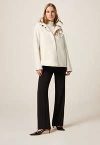 Weiße Kapuzenjacke mit dekorativem Blumenmuster, Knopfakzenten und Vordertaschen. Schwarze, taillierte Hose und glänzende schwarze High-Heels vervollständigen den Look.