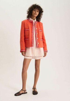 Femme aux cheveux bouclés portant une veste corail texturée avec des boutons dorés sur un chemisier rayé et un short, debout dans des sandales noires tissées.