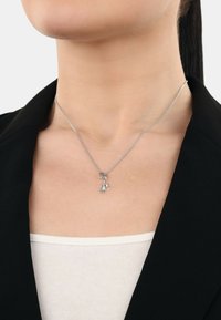 Silberne Halskette mit feiner Kette, die einen kleinen Hund charm in Form eines Hundes trägt, über einem weißen Oberteil und unter einem schwarzen Blazer.