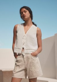 Un top court en lin blanc avec un décolleté en V et une fermeture à nouer, présentant un ourlet agrémenté de pompons ; un short beige avec des motifs floraux brodés.
