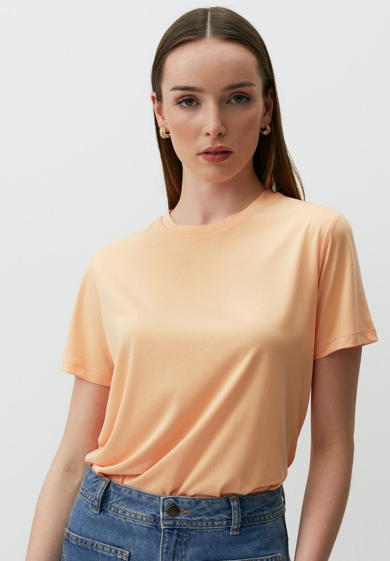 Koszulka z krótkim rękawem, okrągłym dekoltem, uszyta z miękkiego brzoskwiniowego materiału, charakteryzująca się luźnym krojem i gładką fakturą, zestawiona z dżinsami o wysokim stanie.