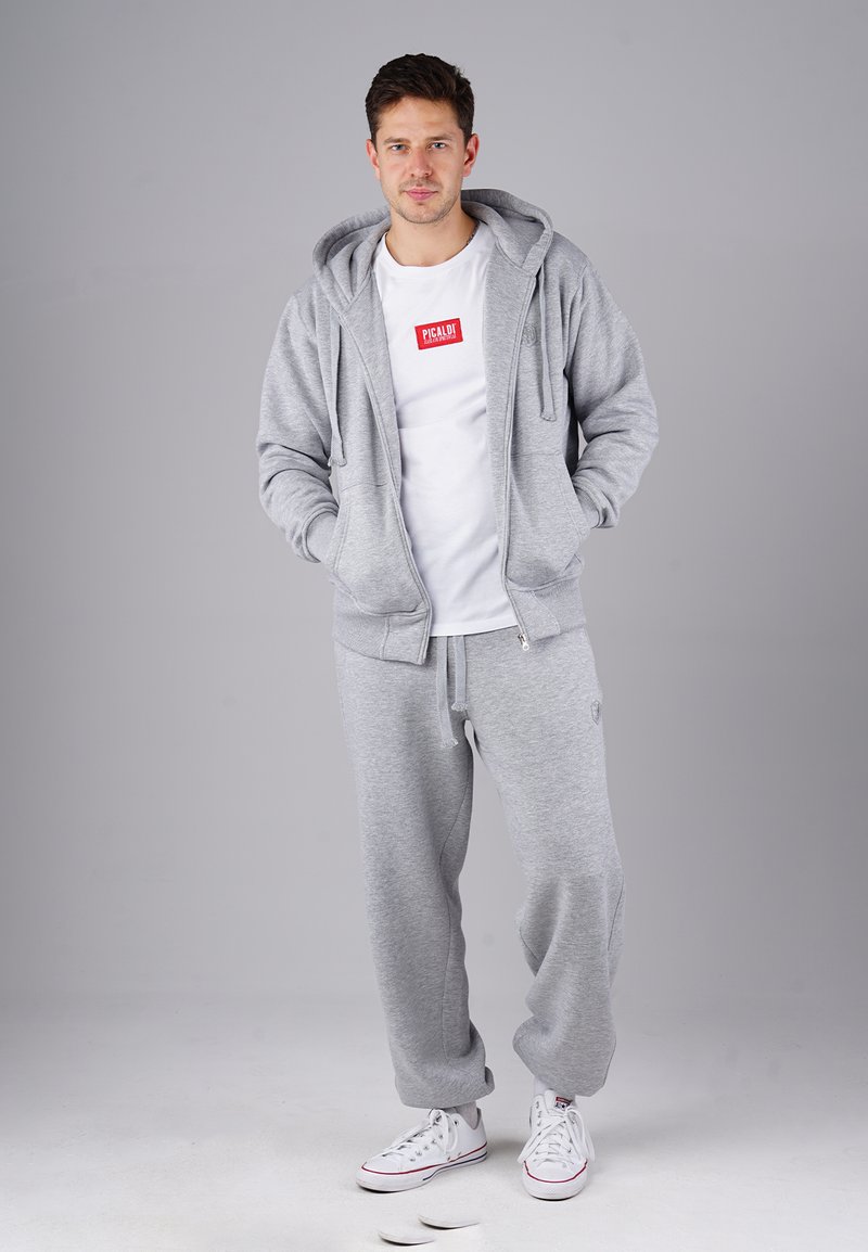 Man wearing a grey zip-up hoodie, matching sweatpants, white sneakers, and a white T-shirt with a red "PICALDI" logo.