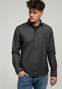 Młody mężczyzna z kręconymi włosami, ubrany w ciemnoszary koszulowy shirt na ciemnym t-shircie i niebieskich dżinsach, patrzy w lewo.