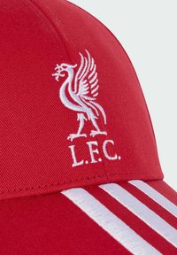 Casquette rouge avec un emblème de merle de Liverpool brodé en blanc et les initiales L.F.C. au-dessus de trois bandes blanches sur le bord.