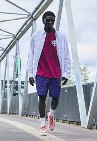 Lilla cargo shorts, en pink grafisk t-shirt, en hvid stribet skjorte med knapper, og pink sneakers. Accessories inkluderer solbriller og en kæde.
