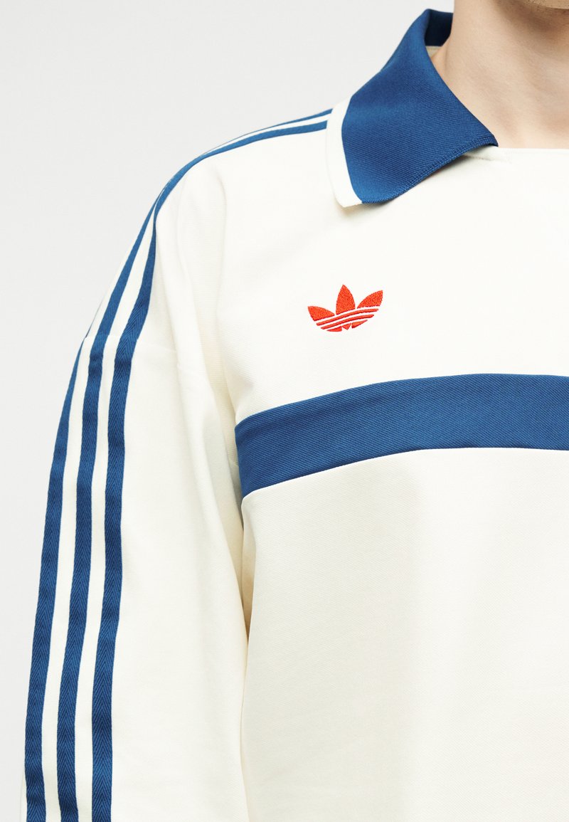 Maglietta bianca a maniche lunghe con strisce blu sulle maniche, fascia orizzontale blu sul petto, colletto blu e piccolo logo rosso sul petto sinistro.