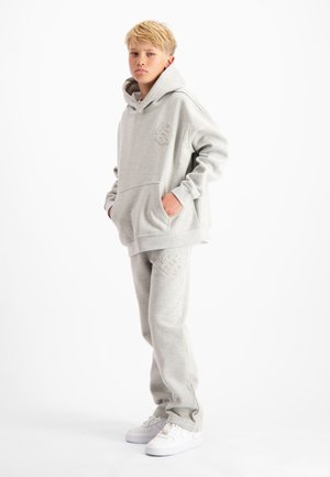 Blonde jongen die met zijn handen in zijn zakken staat, gekleed in een lichtgrijze hoodie en sweatpants met bijpassende logo's en witte sneakers op een witte achtergrond.