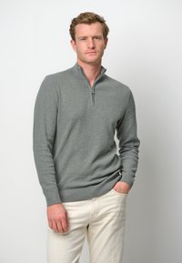 Grauer Zip-Hals-Pullover aus strukturiertem Strickstoff, mit langen Ärmeln und schmalem Schnitt. Kombiniert mit hellen Hosen.