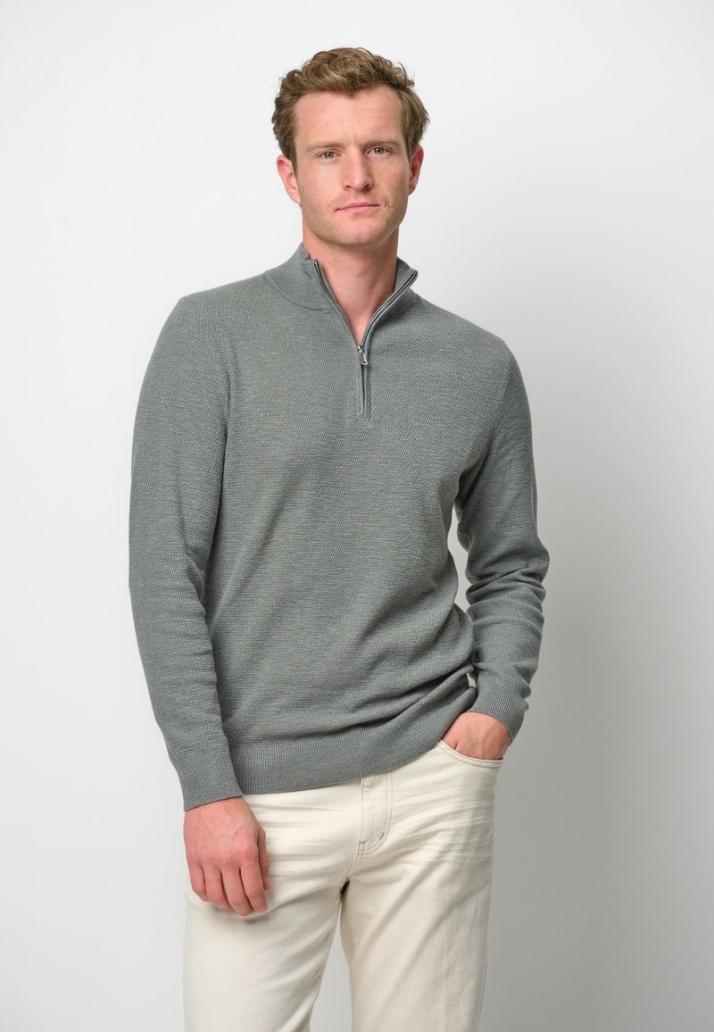 Grauer Zip-Hals-Pullover aus strukturiertem Strickstoff, mit langen Ärmeln und schmalem Schnitt. Kombiniert mit hellen Hosen.