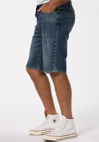 Pantalones cortos de mezclilla en un lavado azul oscuro con un panel más claro en contraste. Presenta presillas para el cinturón y bolsillos frontales. Combinados con zapatillas deportivas blancas de caña alta.