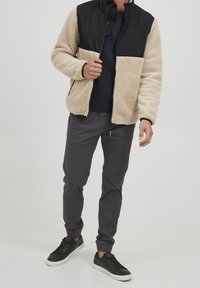 Beige fleece jack met zwart nylon bovenstuk, ritsluiting en twee zijzakken, gecombineerd met grijze taps toelopende broek en zwarte sneakers.
