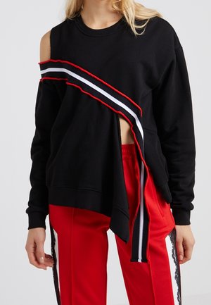 Zwarte sweatshirt met asymmetrische snit, rode en witte gestreepte accenten, en een blootgestelde zijkant. Gecombineerd met rode broek met zijdetails.