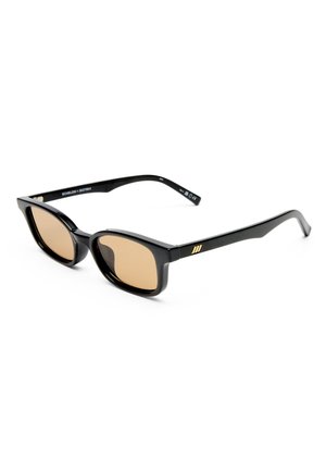 Gafas de sol negras rectangulares con lentes tintados en marrón y un pequeño logotipo dorado en la patilla, descansando sobre una superficie blanca.