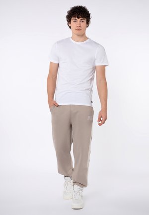 Jonge man met krullend haar die een wit T-shirt, beige joggingbroek en witte sneakers draagt, staand tegen een effen witte achtergrond.