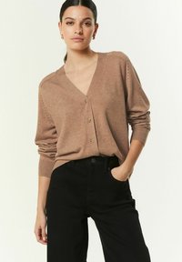 Cardigan tricotat maro cu decolteu în V, închidere cu nasturi și manșete cu model striat. Purtați peste pantaloni negri cu talie înaltă, cu o textură și detalii subtile.
