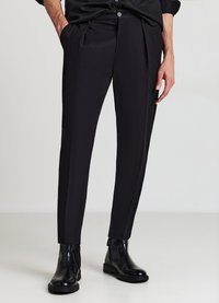 Schwarze, maßgeschneiderte Hose mit glatter Textur, mit seitlichen Taschen und schlankem Schnitt. Kombinierte mit schwarzen Stiefeletten, stehend auf einem hellen Hintergrund.