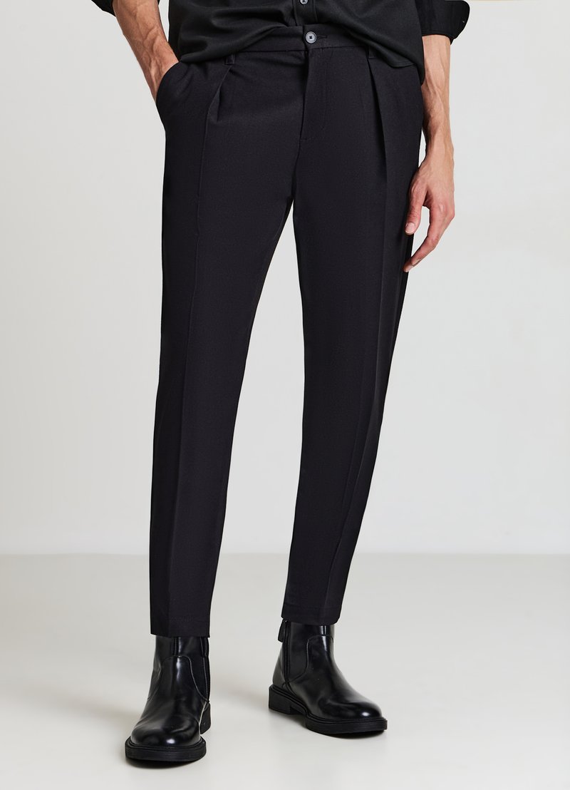 Schwarze, maßgeschneiderte Hose mit glatter Textur, mit seitlichen Taschen und schlankem Schnitt. Kombinierte mit schwarzen Stiefeletten, stehend auf einem hellen Hintergrund.