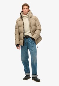 Giacca imbottita beige con texture trapuntata e cappuccio, indossata sopra un maglione chiaro e jeans blu. Una cintura nera e scarpe scure completano l'outfit.