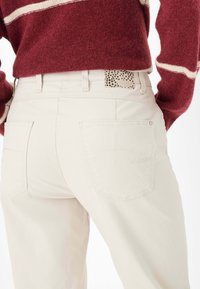 Hellbeige Baumwolljeans mit zwei Gesäßtaschen, die dezente Nähdetails aufweisen. Ein Etikett mit schwarzen Sternen ist am Bund sichtbar.