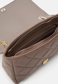 Hnedá prešívaná kabelka s hladkým povrchom, zipsovými vreckami a zlatými kovovými doplnkami. Otvorený vrch odhaľuje podšívku vnútra.
