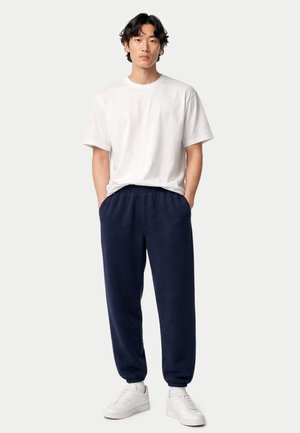 Mann steht mit den Händen in den Taschen einer marineblauen Jogginghose, trägt ein weißes T-Shirt und weiße Sneakers vor einem schlichten hellen Hintergrund.