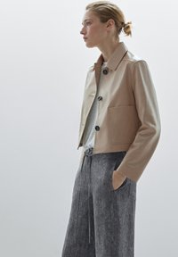 Cropped beige leather jacket with a shirt collar, button closure, and one large pocket; worn over a gray T-shirt and gray trousers.