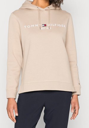 Sweat à capuche beige Tommy Hilfiger avec cordons et texte logo porté avec un pantalon noir sur une personne devant un fond uni.