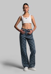 Denim wide-leg jeans with a subtle logo pattern, blue tones, paired with a white ribbed crop top and white sneakers. Silver hoop earrings.