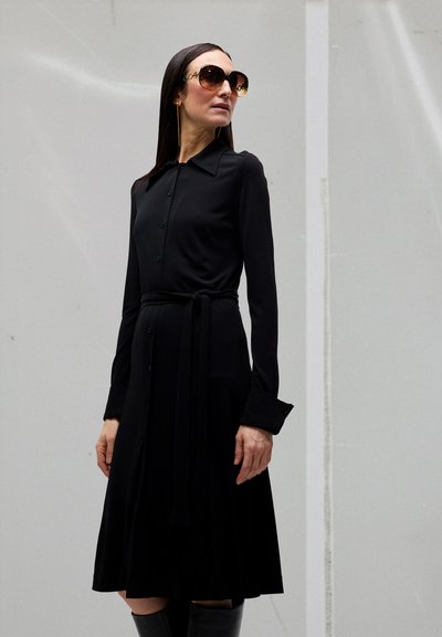 Frau trägt ein langes schwarzes Kleid mit Knöpfen und Taillengürtel, übergroße Sonnenbrille und hat lange glatte Haare, steht vor einer grauen Wand.