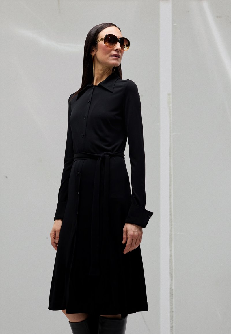 Vrouw draagt een lange zwarte jurk met knopen en een ceintuur in de taille, oversized zonnebril en heeft lang stijl haar, staand tegen een grijze muur.