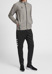 Chaqueta gris con cremallera y bolsillos laterales, combinada con pantalones de chándal negros que tienen rayas blancas y zapatillas negras.