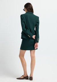 Mujer de pie con la espalda vuelta, vistiendo un blazer y falda verde oscuro, zapatos planos marrones y gafas de sol negras, contra un fondo blanco liso.