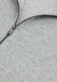 Zbliżenie na częściowo rozpięty srebrny metalowy zamek błyskawiczny na jasnoszarej, teksturowanej tkaninie z delikatnym pionowym żebrowaniem.