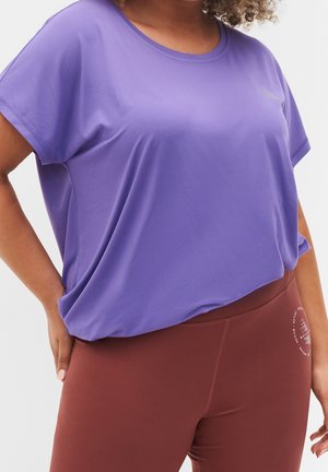 Kobieta w luźnej, fioletowej bluzce z krótkim rękawem i dopasowanych rdzawych legginsach, z ręką opartą na biodrze na białym tle.