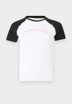 Balta T-krekls ar melnām raglanu piedurknēm un "Guess Jeans" viegli rozā krāsā uzdrukāts uz krūtīm.