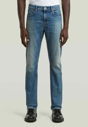 Mann trägt ausgewaschene blaue Jeans und schwarze Schnürstiefel, steht vor einem schlichten hellen Hintergrund.