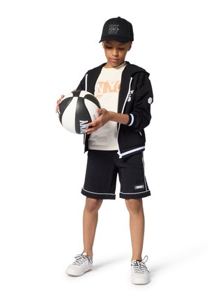 Hoodie noir à fermeture éclair, t-shirt blanc à manches courtes avec texte orange, short noir avec bordure blanche et baskets blanches. Tenant un ballon de basket.