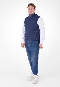 Giacca senza maniche blu navy con colletto alto, chiusura con zip e due tasche laterali. Indossata sopra una camicia bianca a maniche lunghe e jeans blu.