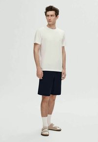 Balta trumpa rankovė t-shirt, tamsiai mėlyni šortai, balti kojinės ir smėliniai sandalai. Paprastas dizainas, lengvas medvilnės audinys. Stovinti poza, neutrali fonas.
