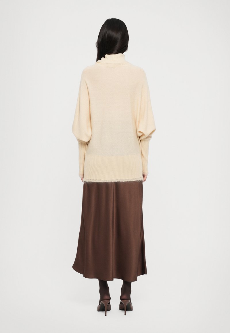 Pull en maille beige avec un col montant et des épaules tombantes, associé à une jupe fluide en satin marron, dotée d'une fente sur le côté et de talons élégants.