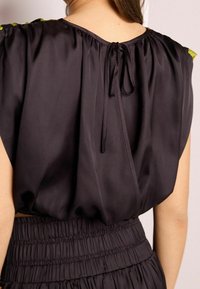 Haut en satin noir avec des épaules froncées et un lien dans le dos, associé à une taille smockée. Texture lisse, coupe décontractée.