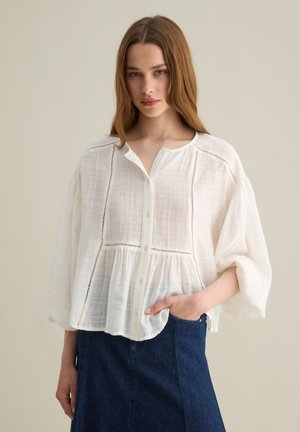 Weiße, lockere Bluse aus einem weichen, strukturierten Stoff, mit einer durchgehenden Knopfleiste und Stickerei-Akzenten. Tragen mit einem Jeansrock.