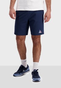 Marinblå idrottsshorts i mjukt tyg; med en broderad logotyp; tillsammans med marinblå sneakers och vita ankelsockor.