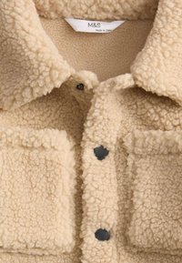 Giacca in pile beige con una finitura testurizzata e soffice, dotata di collo allargato, due tasche anteriori e chiusure con bottoni a pressione neri.